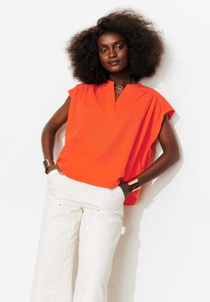 Femme aux cheveux volumineux et bouclés portant un haut sans manches orange vif et un pantalon blanc, debout les mains dans les poches sur fond blanc.