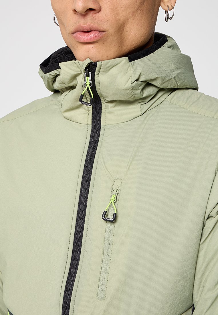 Homme portant une veste à capuche vert clair avec une fermeture éclair noire et des tirettes de fermeture éclair vert fluo, montrant ses lèvres, son menton et ses boucles d'oreilles.