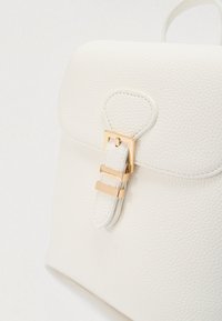 Λευκό σακίδιο πλάτης από δέρμα με υφή, με στρογγυλεμένο καπάκι, πόρπη με χρυσό μεταλλικό στοιχείο και χειρολαβή στην κορυφή για μεταφορά.