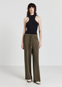 Schwarzer ärmelloser Bodysuit kombiniert mit lockeren, olivgrünen Sweatpants mit elastischem Bündchen und Kordelzug, getragen mit hellen Schuhen.