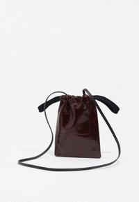 Mała, ciemnobrązowa, błyszcząca skórzana torba na sznurek z cienkim paskiem na ramię i czarną tkaninową rączką na białym tle.