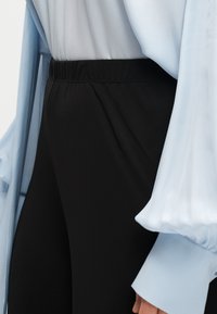 Άτομο που φοράει μαύρο παντελόνι με ελαστική μέση και γαλάζιο μπλουζάκι με μακριά μανίκια φουσκωτά.