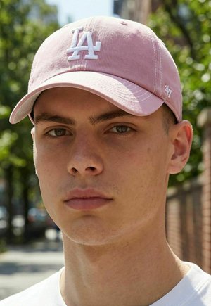Jeune homme portant une casquette de baseball rose LA et une chemise blanche, avec un arrière-plan flou d'extérieur composé d'arbres et de bâtiments.