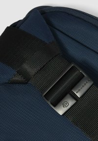 Marineblaue Textiltasche mit einem schwarzen Gurtbandriemen und einer Metallschnalle mit dem "Piquadro"-Logo. Strukturierter Stoff mit diagonalen Rillen.