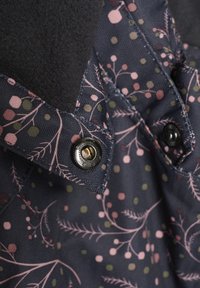 Marinblått tyg med blommönster i rosa och grönt, med svarta knappar och en mjuk fleecekrage; herringbone-textur synlig.