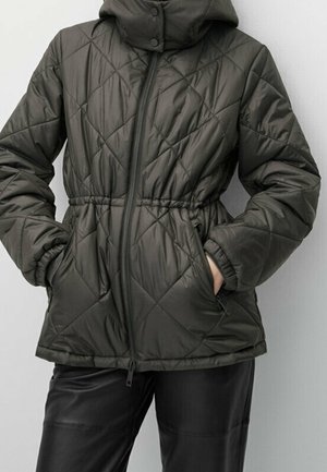 Donkergroene doorgestikte jas met ritssluiting aan de voorkant, elastische taille, hoge kraag, zijzakken, gedragen met een zwarte broek, handen in de zakken.