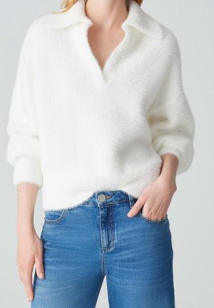 Personne portant un pull blanc pelucheux à col en V et un jean en denim bleu, main dans la poche, devant un fond uni.