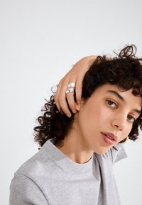 Young person with curly dark hair, wearing a gray shirt, holding their hair back with a hand showing two silver rings.
