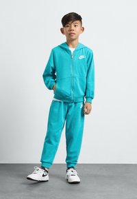 Turkizna Nike kapuca in komplet trenirke z zadrgo spredaj, podrobnostmi žepov in logotipnim poudarkom. Material se zdi mehak in teksturiran. Bele superge.