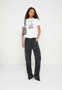 Musta ylisuurikokoinen saharapuku, jossa on useita taskuja, yhdistetty valkoiseen graafiseen T-paitaan, jossa on mustaa tekstiä. Malli kantaa kuvioitua laukkua.