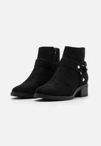 Botines de ante negro con cremallera lateral, presentan una correa con tachuelas plateadas y un tacón bajo en bloque. Textura suave, diseño aerodinámico.