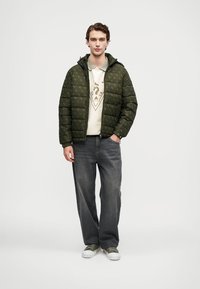 Zielona kurtka puchowa o pikowanym wzorze, noszona na kremowy sweter i szare dżinsy. Model stoi w casualowych trampkach.