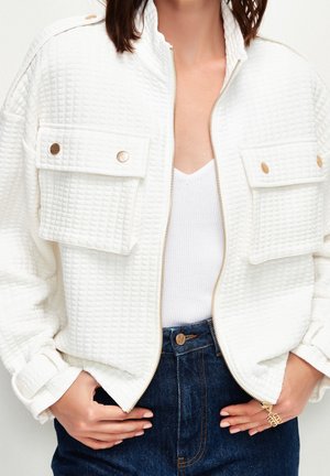 Kvinde iført en hvid tekstureret lynlåstrøje med store klaplommer, en hvid strikket top og blå denimjeans med hænderne i lommerne.