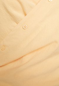 Tecido pêssego claro com uma textura suave, apresentando três botões de cor clara e um prega diagonal, criando um efeito em camadas.