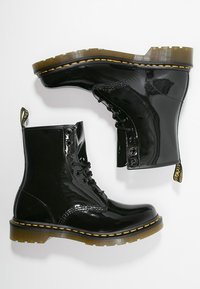 Bottes à lacets en cuir verni noir avec coutures jaunes et semelles épaisses en caoutchouc brun translucide, une chaussure debout et une de côté.