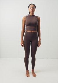 Smeđi bez rukava crop top u kombinaciji s smešnim visoko strukiranim tajicama od glatkog, rastezljivog materijala koji se čvrsto prilagođava telu.