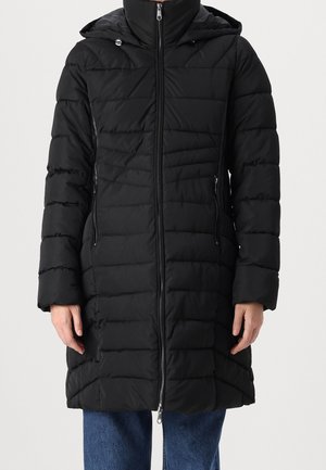 Personne portant un long manteau d'hiver matelassé noir avec capuche, fermeture éclair à l'avant, poches zippées et jeans bleus.