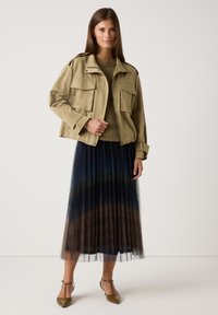 Frau trägt eine beigen, gekürzten Jacke, einen olivfarbenen Strickpullover, einen gefältelten Midi-Rock aus blauer und brauner Tüll sowie olivgrüne spitze Ankle-Strap-Absätze.