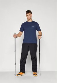 Tumši zila T-krekls ar kabatu, melnas bikses un oranžas pārgājienu kurpes. Modelis pozē ar pārgājienu nūjām pret vienveidīgu fonu.