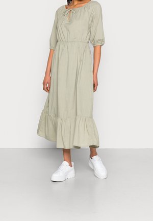 Femme portant une robe midi vert clair avec des manches bouffantes, une taille froncée, un ourlet à volants, et des baskets compensées blanches sur un fond uni.