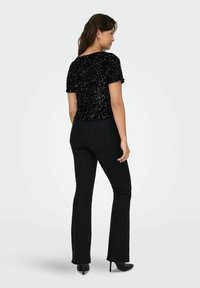 Schwarzes Pailletten-Oberteil mit kurzen Ärmeln, kombiniert mit schwarzen ausgestellten Jeans. Das Oberteil hat eine strukturierte, glänzende Oberfläche und eine figurbetonte Silhouette.