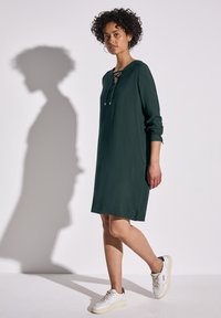 Robe verte jusqu'aux genoux avec un col rond et un détail en lacet. Manches longues avec des poignets. Portée avec des baskets blanches.