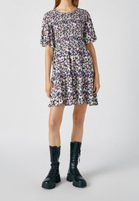 Robe fleurie avec manches courtes volantées, corsage froncé et jupe à volants. Couleurs comprises : violet, crème et vert. Associée à des bottes noires à lacets.