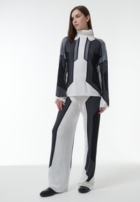 Vrouw in een wit, zwart en grijs geometrisch patroon coltrui en bijpassende wijde broek met zwarte schoenen, staande voor een witte achtergrond.