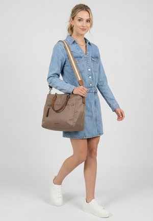 Blondine in een spijkerjurk die loopt, met een taupekleurige handtas met tan- en beige band, witte sneakers dragend op een grijze achtergrond.