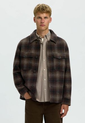Ung mand iført en brun ternet jakke med knapper over en beige skjorte, stående med den ene hånd i lommen mod en lys ensfarvet baggrund.