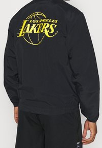 Schwarze Windjacke mit großem gelben "Los Angeles Lakers"-Logo und Basketballgrafik auf dem Rücken.