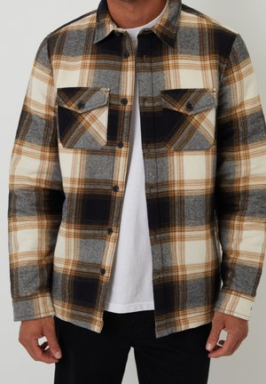 Homme portant une chemise à carreaux marron, noire, grise et crème boutonnée sur un t-shirt blanc avec un pantalon noir, debout devant un fond uni.