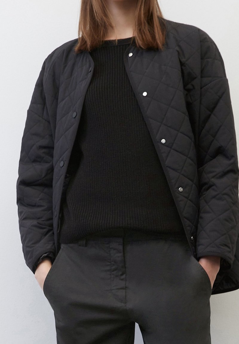 Personne portant une veste matelassée noire par-dessus un pull en tricot noir et un pantalon noir, les mains dans les poches, sur un fond clair uni.