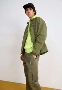 Ελιά πράσινο μπουφάν με μαύρες λεπτομέρειες, νεον πράσινη κουκούλα και ελιά cargo παντελόνι. Το σύνολο έχει χαλαρή γραμμή και εμφανή λογότυπα.