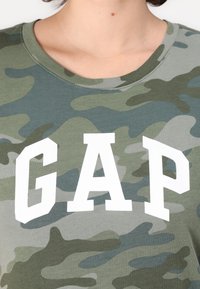 Grønn kamuflasjemønstret t-skjorte med hvit "GAP" trykk. Skjorten har rund hals og er laget av myk, lett bomullstoff.