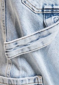 Lys blå denimstoff har en lomme og oppbrettede ermer. Teksturen viser klassisk indigo-fading og kontrasterende oransje sømdetaljer.