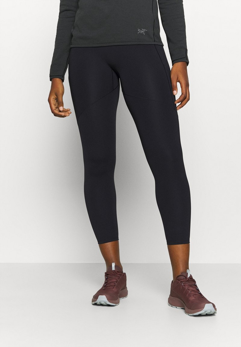 Personne portant des leggings noirs courts et des chaussures de sport bordeaux, debout sur un fond blanc.