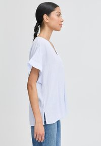 Maglietta oversize a righe bianche con scollo a V, maniche corte e spacchi laterali, abbinata a jeans blu. Texture liscia, design casual.