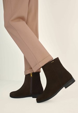 Osoba w beżowych spodniach i ciemno-brązowych zamszowych botkach za kostkę z bocznym zamkiem i niską czarną podeszwą, stojąca na jasnej powierzchni.