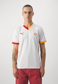 Maillot de football blanc avec un col en V, accents rouges et jaunes sur le col et les manches, comportant un logo et du texte, ainsi qu'un motif texturé.