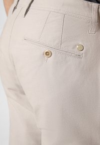 Beige trousers made of textured fabric, featuring a back pocket with a button closure and a small metal accent.