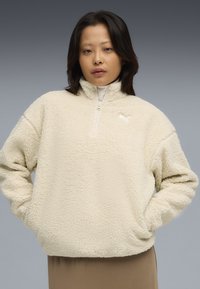 Cremefarvet fleece-pullover med høj krave, halv lynlås og logoaccent på brystet. Blød, struktureret overflade.