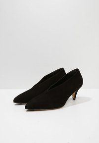 Chaussures basses en daim noir avec bouts pointus et ouvertures en forme de V. Les talons mesurent environ 5 cm de hauteur, offrant une silhouette élégante.