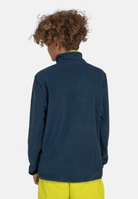 Giacca in pile blu navy con colletto alto, caratterizzata da una texture liscia e maniche lunghe, abbinata a pantaloni gialli brillanti.
