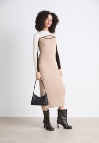 Strickkleid in Beigetönen, Creme und braunen Farbkombinationen mit langen Ärmeln und einem runden Ausschnitt. Schwarze Handtasche und glänzende schwarze High-Heels.