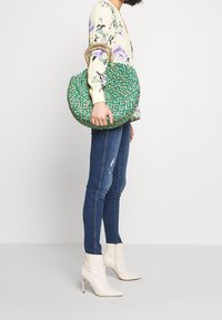 Green woven handbag with a round shape and natural rope handles, featuring a textured pattern. Model holding the bag while wearing jeans and white boots.