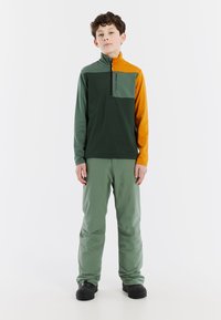 Maglia con zip verde e arancione, con tasca sul petto, abbinata a pantaloni verde chiaro e calzature nere. Tessuto morbido e resistente, vestibilità atletica.