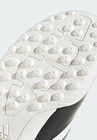 Gros plan de la semelle d'une chaussure de sport blanche avec des crampons en caoutchouc ronds et un motif de grip texturé sur une base noire et blanche.