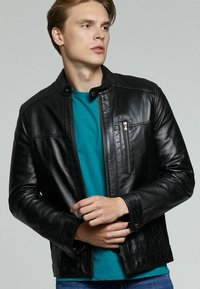Chaqueta de cuero negra con collar alto, bolsillos con cremallera y una textura suave. Llevada sobre una camiseta azul verdosa, mostrando un diseño ajustado.
