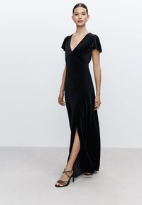 Abito lungo in velluto nero con scollo a V, maniche a svolazzo e spacco laterale. Abbinato a sandali neri con tacco alto e cinturini.